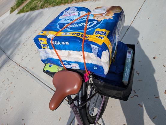 A photo looking down at the back half of a bicycle, parked on a patch of concrete. The bicycle has two fold-down cargo baskets on either side of the rear wheel, both of which are filled with groceries. The one on the right also has a library book (The Water Outlaws by S. L. Huang) tucked in next to the reusable canvas grocery bags. Spanning between the two cargo baskets is a large bulk pack of toilet paper, secured with bungee cords. The visible part of the bicycle has a brown seat and a light purple frame.