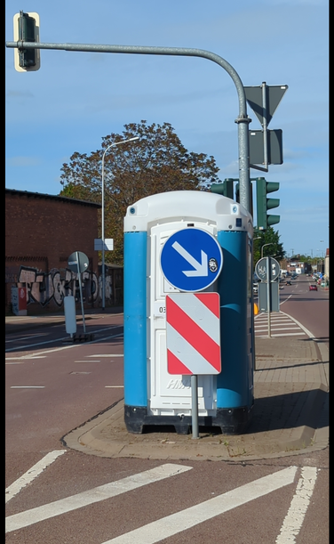 auf eine Mittelinsel steht hinter der Richtungsbeschilderung ein ToiToi Toilettenhäuschen.
