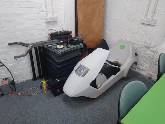 A picture of the Sinclair C5 showing it fully disassembled in a corner, with the plastic body sitting on the floor surrounded by it's components.