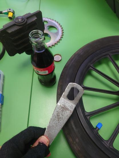 A picture of me holding the flat cone spanner next to an open glass bottle of cola, on a table with the pedal crank and sprocket and rear wheel.