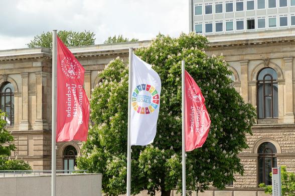 Die Flagge der UN-Nachhaltigkeitsziele weht zwischen zwei TU-Fahnen. Im Hintergrund ist das Altgebäude der TU Braunschweig zu sehen.

Foto: Kristina Rottig/TU Braunschweig