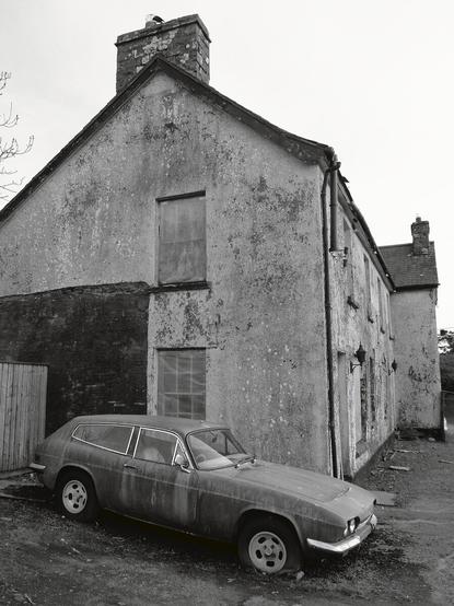An old car parked, apparently for a very long time, behind a house as abandoned as it seems to be