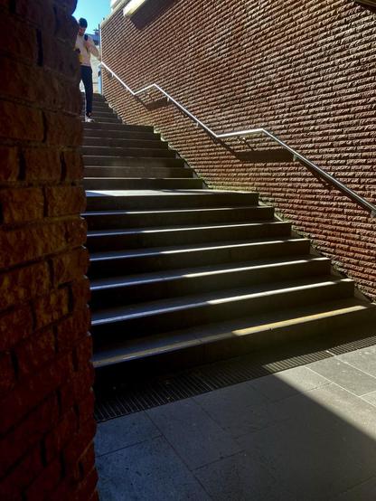 Zu sehen ist die linke Treppe des hinteren Ausgangs der Bonner Bahnhofspassage. Die Treppe hat zwei Podeste, deren Absatz sich im Geländer als gerades Teilstück wiederholt. Von oben scheint die Sonne hinunter. Eine Person ist ganz oben auf der Treppe zu sehen, die hinuntergeht. Daneben erstrahlt ein winziges Stück blauer Himmel. Die Sonne sorgt für einen soliden Schattenwurf.
