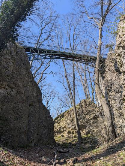A metal bridge with wooden planks for hikers connecting the top of two large rocks viewed from underneath.