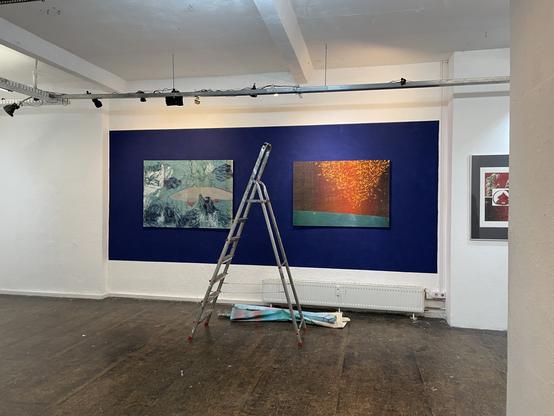 Ausstellungsaufbau. Zwei großformatige Linolschnitte hängen an einem blauen Wand. Ein Leiter in der Mitte.