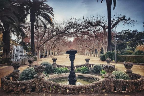 A stone fountain sits in the foreground of a park, with a long, tree-lined walkway stretching into the distance under a cloudy sky.