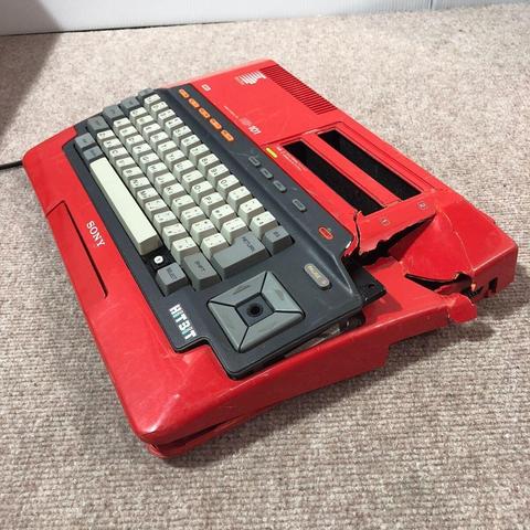 a red and grey Sony HiTBiT HB-101 personal computer. It features a QWERTY keyboard with white keycaps and a darker grey function key area to the right, including cursor keys, RETURN, STOP, and PAUSE buttons. The "HiTBiT" logo is visible in white lettering on a grey background to the left of the cursor keys. The "SONY" logo is embossed in white on the red casing to the left of the keyboard.

The computer has slots for two cartridges on the right side, labeled "1" and "2". There are also several red buttons and indicator lights located above the keyboard on the right side of the main unit. The casing appears to be made of plastic and shows some signs of wear and tear, particularly on the right side where some parts look cracked or broken. A black power cable is visible extending from the left side of the computer.