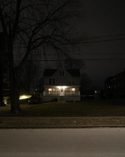 Color photograph looking across a brightly lit street at night at a large two-story house with an attic set quite a ways back behind a big flat front lawn, a large tree up close by the sidewalk is silhouetted against the glowing overcast sky