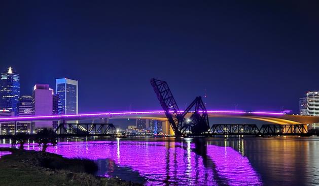 Overnight view of the colorful downtown Jacksonville skyline and bridges. Beneath a clear, dark blue sky, the building and bridge lighting illuminate the night sky, and the Acosta Bridge's neon stripe casts purple reflections across the water's surface below.