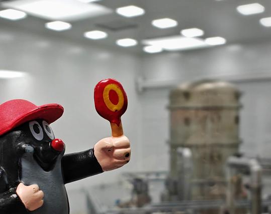 A close-up of the 'small mole' toy with a red cap and red nose, holding up a red and yellow lollipop. The background shows a blurred laboratory setting with ceiling lights and vacuum vessels.