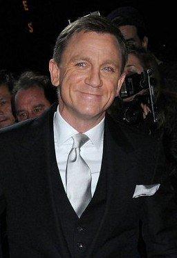 Daniel Craig lächelnd in formeller Kleidung. Er steht in einer Menschenmenge und blickt direkt in die Kamera. Im Hintergrund sind unscharfe Gesichter und Kameras zu sehen.