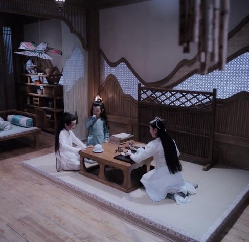 Three children kneel at a table - Wei Ying and Jiang Cheng beside each other on one end and Jiang Yanli at the other. A white bowl with a lid sits in front of Wei Ying, and it's twin sits on a black tray in front of Yanli along with two spoons. Yanli is shown mostly from behind, but enough is visible of her profile to tell that she is smiling at the boys. Wei Ying also sports a bashful smile while looking down at the bowl in front of him, and Jiang Cheng looks on expectantly.