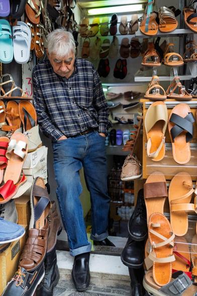 A candid photo taken in a small, crowded shoe shop in Athens, Greece, showing an older man with grey hair wearing a plaid shirt and blue jeans, leaning casually in the doorway surrounded by a colourful and diverse array of sandals, slippers, and shoes displayed on shelves and boxes. Some of the visible shoe brands include "Sabio" on a pair of white and blue sandals.