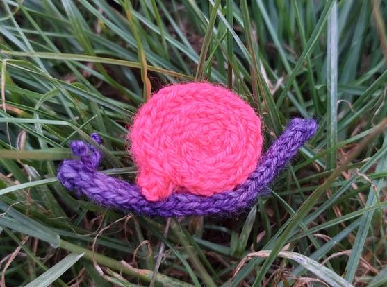 A very small knitted snail, with a purple body and bright pink shell, lying in grass.