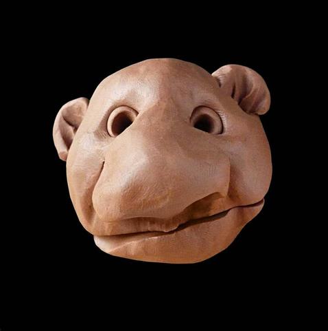 The image shows a sculptural head of what appears to be a cartoonish or stylized bear or bear-like character. It's made from a clay or terracotta-colored material with a smooth, polished surface. The sculpture features a rounded head with prominent circular eye sockets, a broad snout with nostrils, large ears, and a slightly open mouth that gives it a gentle, somewhat friendly expression. The head is isolated against a solid black background which makes the warm, reddish-brown color of the sculpture stand out dramatically. The craftsmanship appears to be quite detailed, with subtle textures and contours that give the character a distinctive personality.