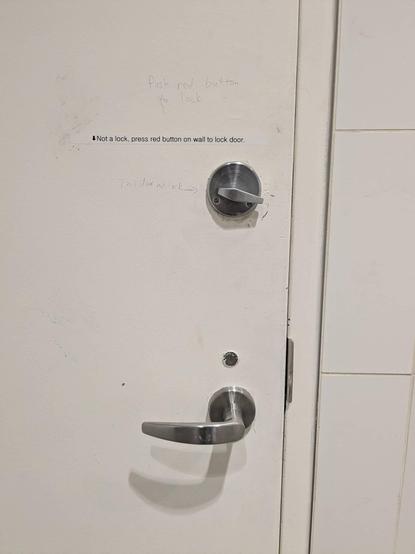 Fairly tightly cropped photo of part of a white door, door frame, and white tiled wall, showing the metal door handle and above it what looks like a standard thumb-operated rotating door latch.

Immediately above the latch is a sign printed with a label maker that says "⬇️ Not a lock, press red button on wall to lock door.", above that a handwritten message on the door says "Push red button to lock" and to the left of the latch another handwritten message says "This door not lock" with an arrow pointing to the latch.

No red button is visible in the photo.