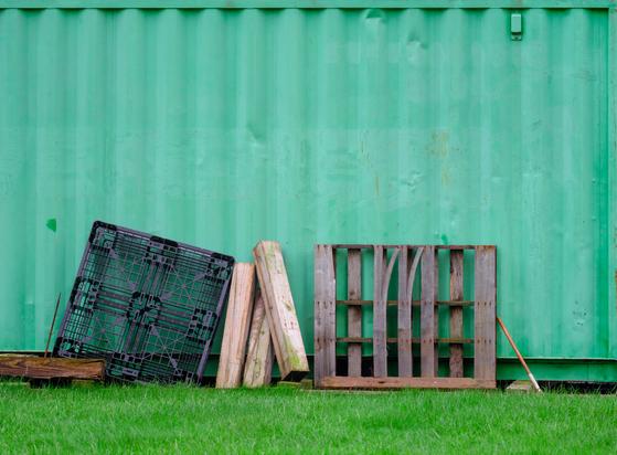 We see multiple colors (aged and gray, yellowish, darker) of wood in multiple forms leaning against the wall of a laarge metal construction waste container painted a delightful shade of greenish.  There's also a black plastic thingie, and there's grass in the foreground.  The colors and textures (but especially the colors) are lovely.