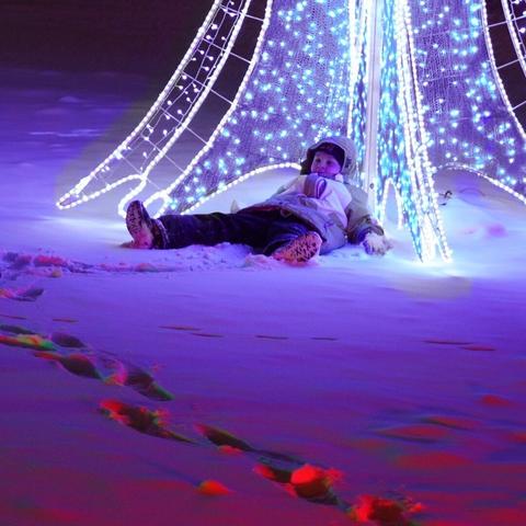 Photograph of a young child slumped in the snow, sitting with his back to a Christmas light display.