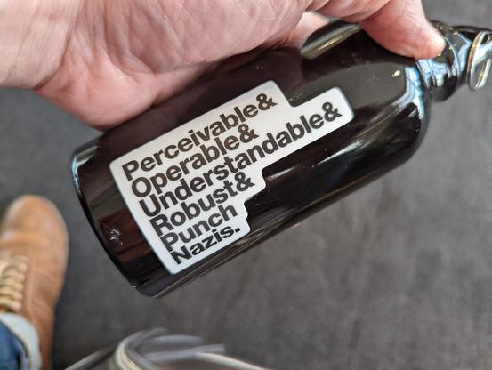 A short black metal water bottle with a sticker of black text on white reading, “perceivable & operable & understandable & robust & punch nazis.”