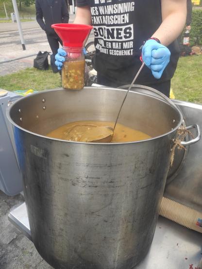 Wielki gar z zupą, z którego jest ona nakładana do słoika z czerwonym lejkiem.