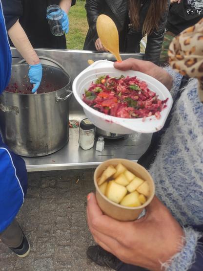 Po prawej stronie widać ręce trzymająca sałatkę i kompot, a w tle osobę nakładającą kolejne danie z wielkiego garnka.