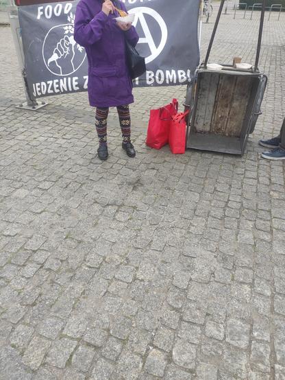 Postać stojąca na tle baneru Food Not Bombs w trakcie spożywania swojego posiłku