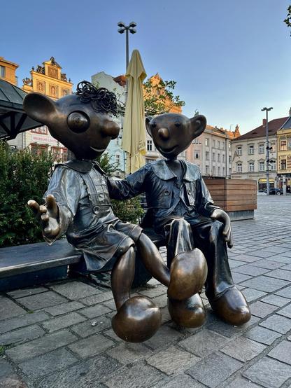 Das Bild zeigt zwei bronzene Figuren, die auf einer Bank sitzen und sich unterhalten, vor dem Hintergrund einer europäischen Stadtlandschaft.


Detaillierte Beschreibung:

Das Foto zeigt eine Nahaufnahme von zwei bronzenen Skulpturen, die auf einer dunklen Holzbank sitzen. Die Figuren sind stilisiert und erinnern an Figuren aus einem Kinderbuch oder Zeichentrickfilm.  Sie haben große Köpfe mit großen Ohren, großen Augen und kleinen, leicht nach oben gebogenen Nasen.  Ihre Körper sind im Vergleich zu ihren Köpfen klein und wirken etwas unbeholfen. Die Figur links hat krauses, dunkles Haar.  Beide tragen einfache, etwas lose Kleidung –  die Figur links scheint ein Jackett und einen Rock zu tragen, während die Figur rechts ein Jackett und Hosen trägt. Beide Figuren haben große Füße, die wie große, runde Klötze aussehen. Die Figur links streckt eine Hand aus, als wolle sie etwas zeigen oder jemanden begrüßen. Die beiden Figuren scheinen sich in einem vertrauten und freundlichen Gespräch zu befinden.  Ihre Körperhaltung und die Art, wie sie sich berühren, strahlen Wärme und Zuneigung aus.

Im Hintergrund ist ein Ausschnitt einer europäischen Stadt zu sehen.  Die Gebäude sind hell und haben eine klassische Architektur.  Ein Teil eines gelben Sonnenschirms ist zwischen den Figuren und den Gebäuden sichtbar.  Vor der Bank sind grüne Büsche zu sehen.  Der Boden besteht aus grauen Steinplatten. Die allgemeine Atmosphäre ist friedlich, ruhig und einladend.