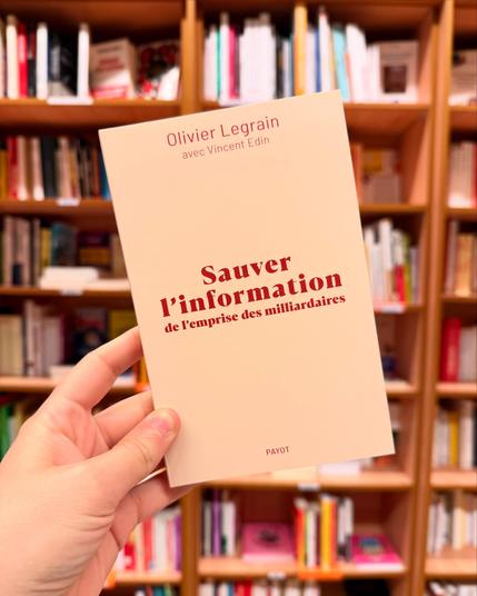 Livre pris en photo devant une bibliothèque de livres. La couverture du livre est tout simple: titre et nom d’auteur en rouge sur fond beige.