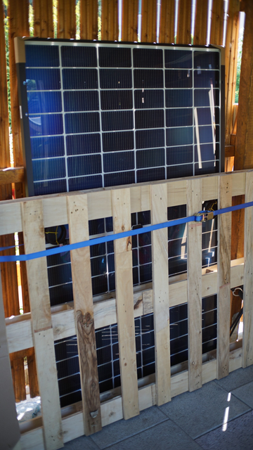 Ein Solarpanel ist an einer Holzwand angebunden (Palette steht davor, Spanngurt um die Palette, damit flächiger Druck auf das Panel)