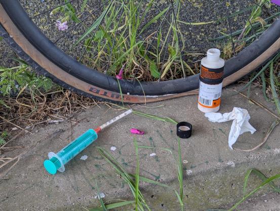 a bicycle wheel with a bottle of sealant, a syringe, a piece of paper towel, and a valve cap, all sitting on a dusty concrete. the tire is deflated, and there are traces of white liquid on the ground and the wheel