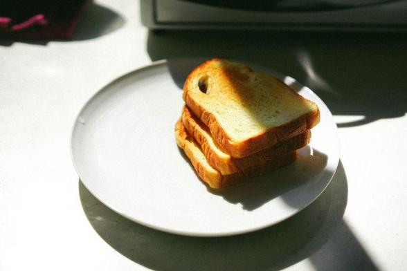 A stack of three pieces of toast on a white plate, under the shadow of the toaster in bright sunlight.