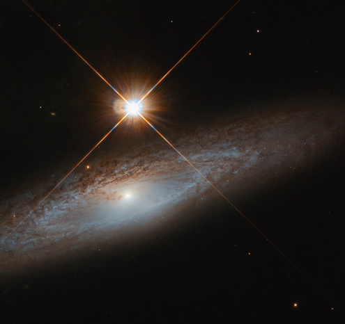 A bright foreground star dominates the upper portion of the frame with diffraction spikes, while the galaxy UGC 3855 stretches across the bottom with a luminous core and swirling bands of dust and bluish star clusters. The black background is dotted with tiny stars and distant galaxies.