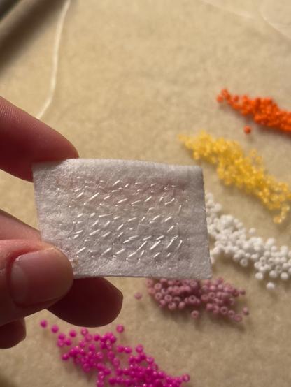Showing the back of my pellon before trimming the excess and adding the backing and edging. You can see all my stitches filling the shape of a flag (rectangle). In the background you can see the beads used--magenta, mauve, white, yellow-orange, and orange-- the lesbian pride colors.