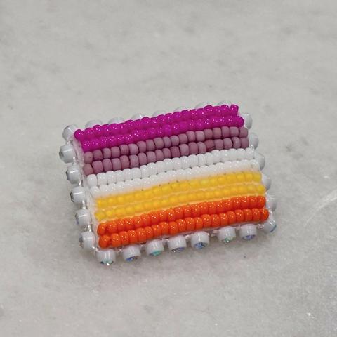A beaded lesbian pride flag pin edged with glass rhinestone banding on a white marble background