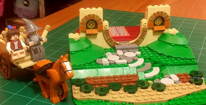 An unfinished Lego build. Gandalf and Frodo are riding in a horse-drawn cart, next to a half-built Hobbit-hole house in a hill that hasn't been built yet.