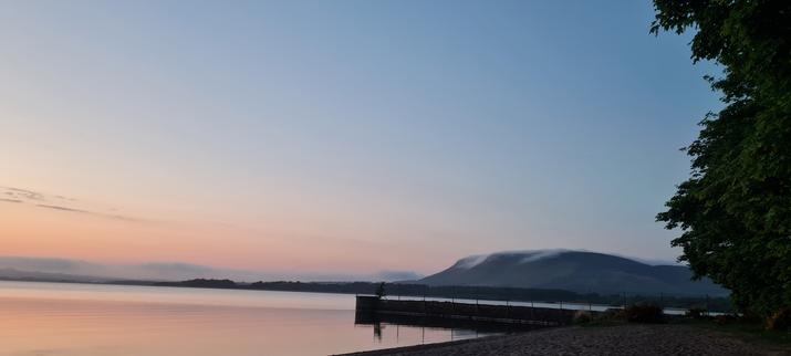 In the distance to the right is a hill with wisps of cloud skirting the top. The sky is blue and changes from gold, then pink towards the left of the picture.

A small wall starts on the right of the beach, near some trees, and stretches into the Loch, stopping in the centre of the picture.

To the left is a view of the still water of the Loch.