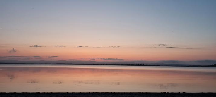 A view of the Loch looking west as the sun has set.
There is a thin layer of purple cloud along the horizon, fading to pink, gold, and blue towards the top of the image.

The Loch water is still and reflects the sky.
