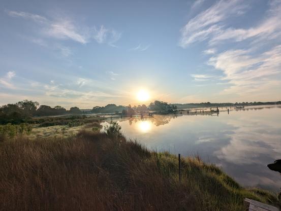 Photo of a calm morning sunrise over Harris Creek sun rising low to the right in a pale blue sky. Wispy white clouds decorate the center and horizon . Marsh is green with riotous new spring growth. Creek water is rippled and reflects the sun, sky and clouds.