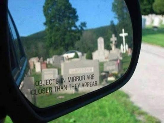 Photo of a side mirror on a car. 

A graveyard full of headstones can be in the rear vision mirror.  

Text on the mirror reads: OBJECTS IN MIRROR ARE CLOSER THAN THEY APPEAR