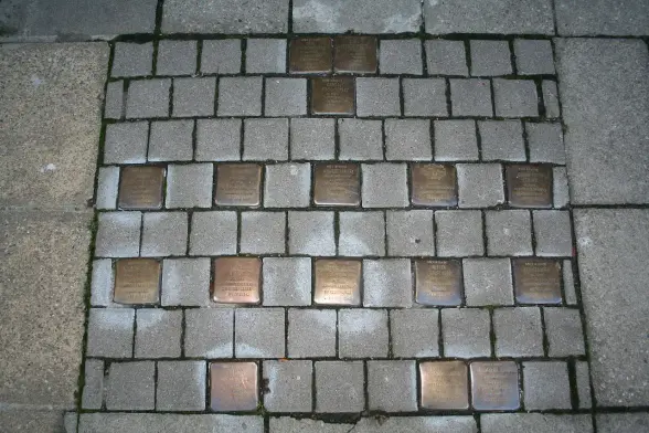 The image shows a street or pavement with embedded plates or plaques. These plaques usually have inscriptions, often commemorating individuals or events. They are arranged in a grid-like pattern on the surface of the pathway. The context of the plaques could vary, but similar plaques are commonly associated with memorials or historical significance, such as honoring victims of persecution.

Image Credits: Wikimedia / Einsamer Schütze / CC BY-SA 3.0