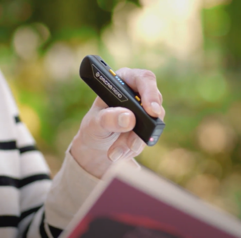 Close-up view of the OrCam Read 5 being held by a woman in her left hand. The reader is black with yellow buttons and a series of blue LEDs on the top of the device. She is pointing the reader slightly downward toward the page of an open book. Her index finger is pushing a button on the top of the reader. A couple additional buttons on top of the device and a red LED on the end of the reader pointed at the book can be seen. The company name ORCAM and the device name READ are visible on the side of the reader.