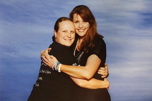 Two people posing for a camera, while embraced in a big hug. Both are smiling. I am on the left, a slightly overweight person, dressed in black. Amanda Tapping is on the right, slender and beautiful, dressed in black as well.