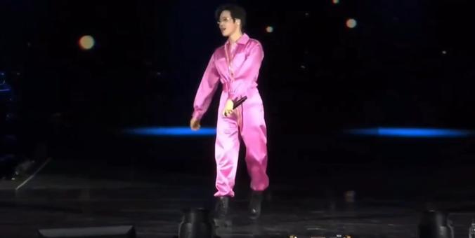 Kpop star SUHO in a pink satin jumpsuit on stage holding a microphone in one hand. He is mid stride and looking to his right.