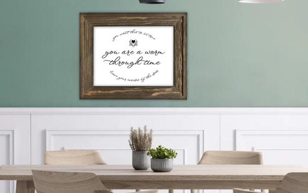 Dining room stock photo featuring a light, natural wood table with two small houseplants on it and matching chairs. The wall behind the table is white with decorative moulding, then halfway up, the wall becomes flat and green. A brown wooden picture frame on the wall contains the focus of the image, fancy black cursive lettering on a white background. The center, in large print, reads "you are a worm through time". Above it is a heart with lines splaying outward from it and a line of text, curved upward, which reads "you want this to be true". Below, a line of text curved downward reads "leave your insides by the door".