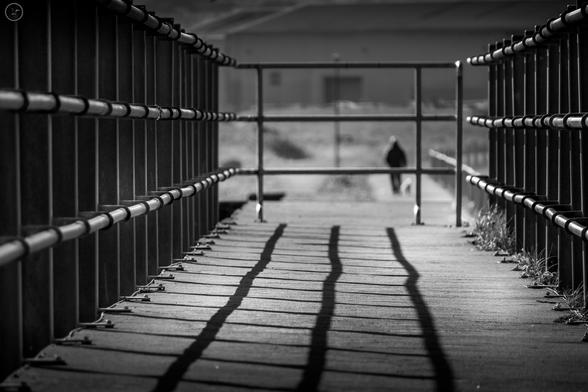 Railings either side of a path casting shadows onto the concrete path. Walker in far distance