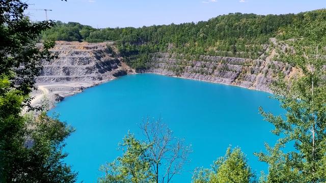 Foto von oben in einen Steinbruch. Das Bild ist eingerahmt von grünem Bewuchs. Die Grube ist terrassiert und es sind ca. 7 Stufen zu sehen. Auf der linken Seite sieht es nach aktivem Bergbau aus, rechts sind die Stufen bereits wieder bewachsen. Mittig nimmt den grössten Teil des Bildes serh braues Wasser auf dem Grund der Grube ein. Am oberen Bildrand ist etwas Himmel zu sehen der im Vergleich zum Wasser eher blass daherkommt.