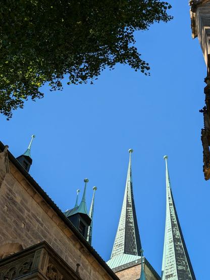 von unten an einer Kirche in den blauen Himmel fotografiert.
6 spitze Turmspitzen mit kleinen Kugeln drauf und grünlichem Kupferspan sind zu sehen