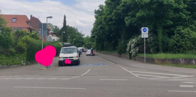 Fahrradstraße mit am "Fahrbahnrand" abgestellten KFZ. Anderes KFZ sucht im Schritttempo einen Parkplatz. Rollerfahrer und Fahrradfahrer sind ebenso gezwungen Schritttempo zu fahren bzw. im Gegenverkehr bis zum Stillstand abzubremsen. Ein VW-Bus steht fast an der Kreuzung auf dem Riesen-Piktogramm "Fahrradstraße".
