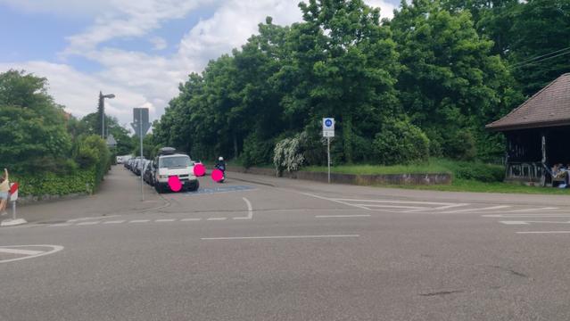 Fahrradstraße mit am "Fahrbahnrand" abgestellten KFZ. Anderes KFZ sucht im Schritttempo einen Parkplatz. Rollerfahrer und Fahrradfahrer sind ebenso gezwungen Schritttempo zu fahren bzw. im Gegenverkehr bis zum Stillstand abzubremsen. Ein VW-Bus steht fast an der Kreuzung auf dem Riesen-Piktogramm "Fahrradstraße".
