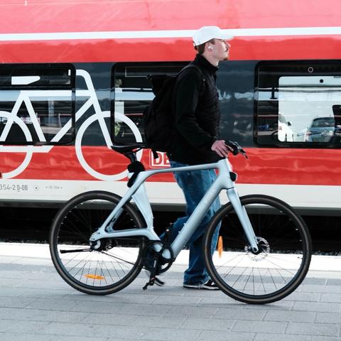 Ein junger Mann schiebt ein helles Pedelec auf einem Bahnsteig vor einem roten Wagon mit einem großen weißen Fahrrad-Piktogramm. Er trägt eine weiße Baseballkappe, eine schwarze Jacke, blaue Jeans und Turnschuhe. Er trägt einen schwarzen Rucksack und weiße Ohrhörer. Er läuft links von seinem Fahrrad - vom Betrachter aus dahinter. Das Fahrrad ist ein Pedelec mit moderner Rahmenform, ohne Schutzbleche und mit recht breiten Reifen. Im Hintergrund befindet sich ein roter Zug, auf dessen Seite ein großes weißes stilisiertes Fahrrad abgebildet ist. 

A young man pushes a light-colored pedelec on a platform in front of a red wagon with a large white bicycle pictogram. He is wearing a white baseball cap, a black jacket, blue jeans and sneakers. He is carrying a black rucksack and white earphones. He is walking to the left of his bike - behind it from the viewer's perspective. The bike is a pedelec with a modern frame, no mudguards and fairly wide tires. In the background is a red train with a large white stylized bicycle on the side.